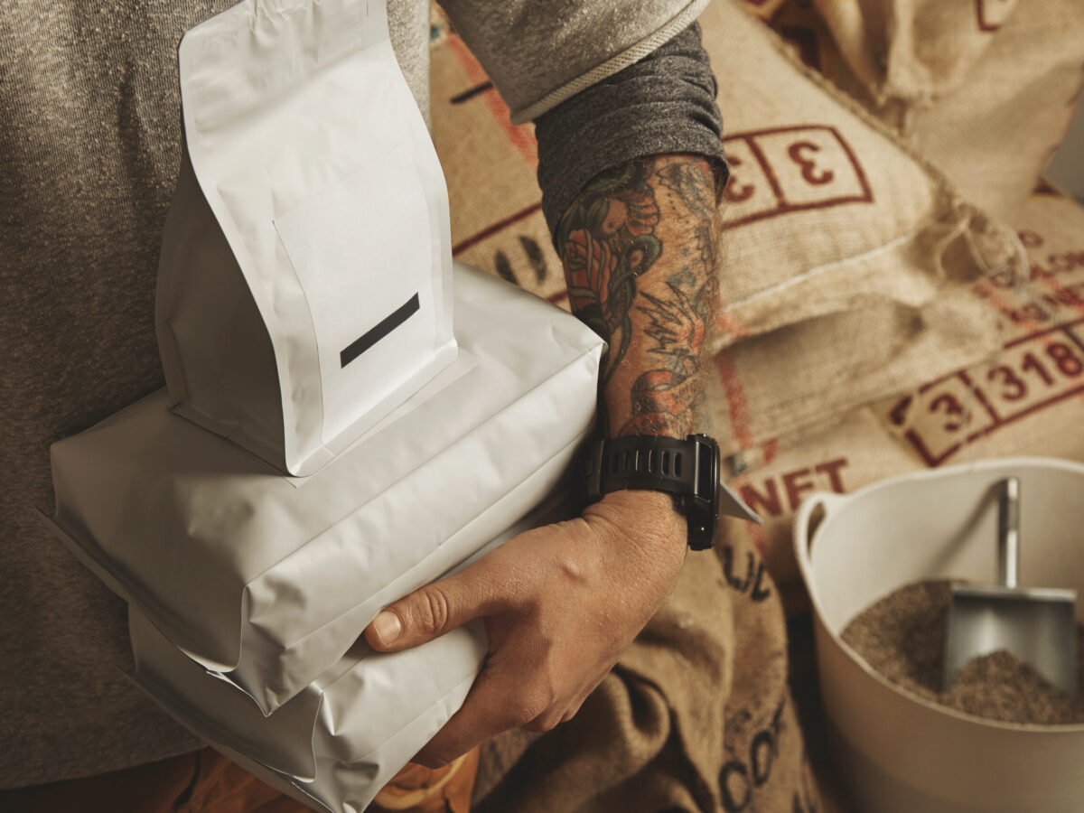 Tattooed barista holds blank package bags with freshly baked coffee beans ready for sale and delivery Plastic basket near big bags on europalet in warehouse behind Professional alternative roasting and brewing